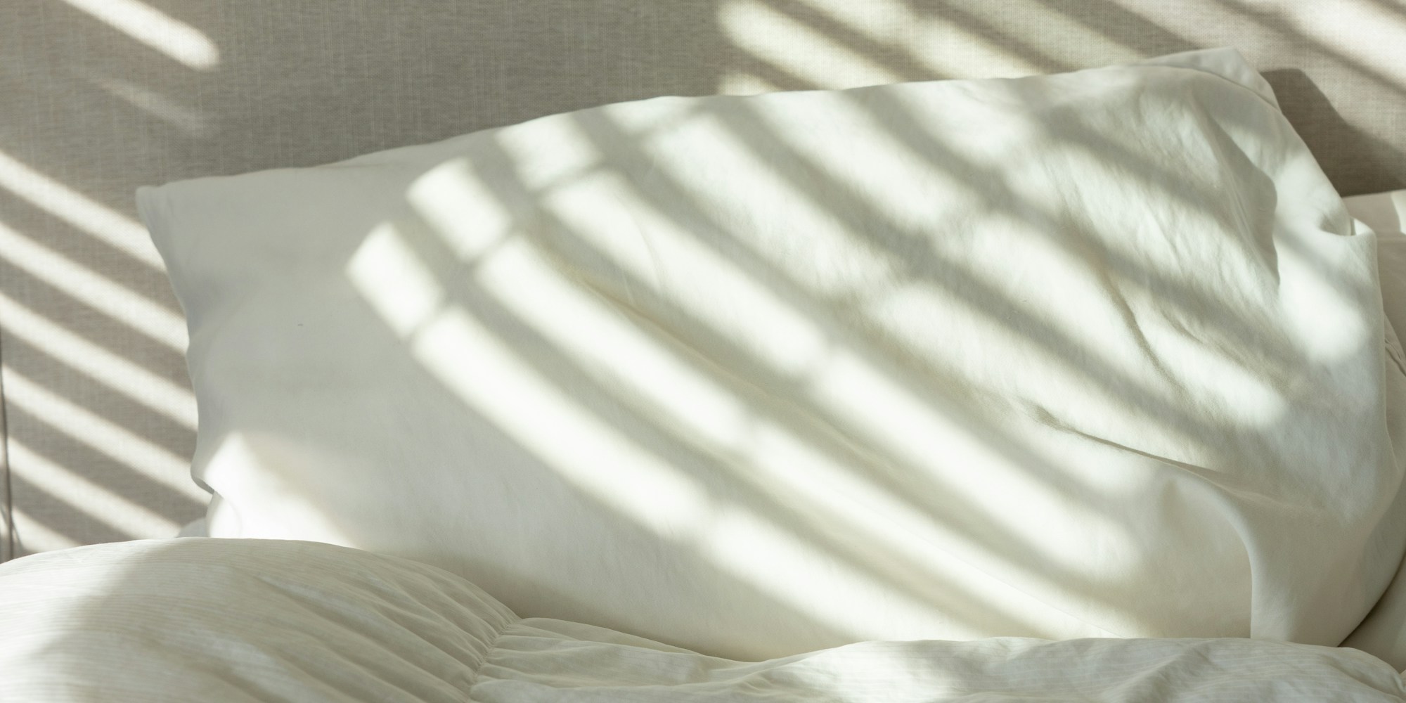 Un oreiller blanc sur un lit clair, traversé par la lumière rayée de persiennes, format panoramique