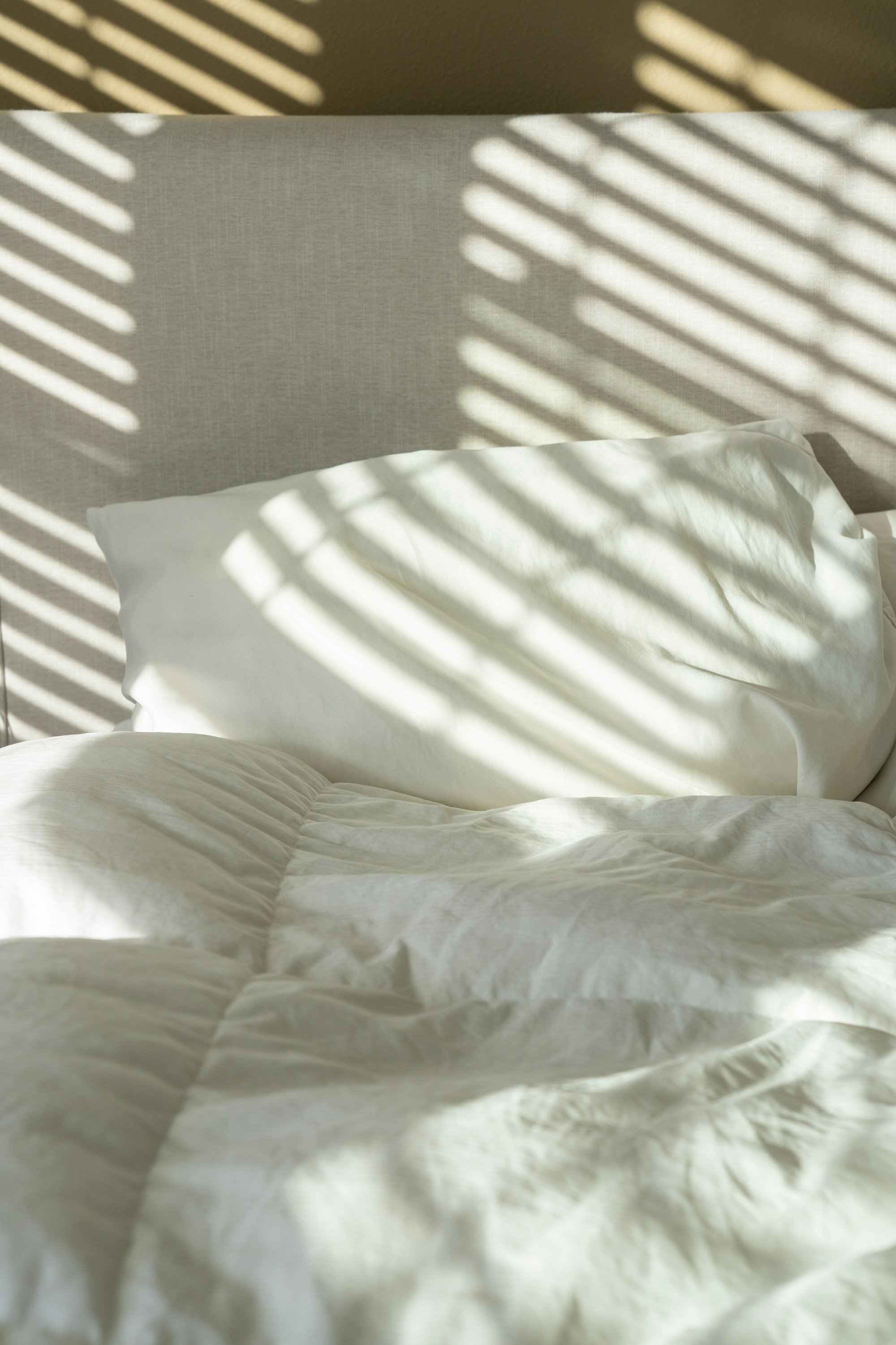 Un oreiller blanc et une couette froissée, traversés par la lumière rayée de persiennes